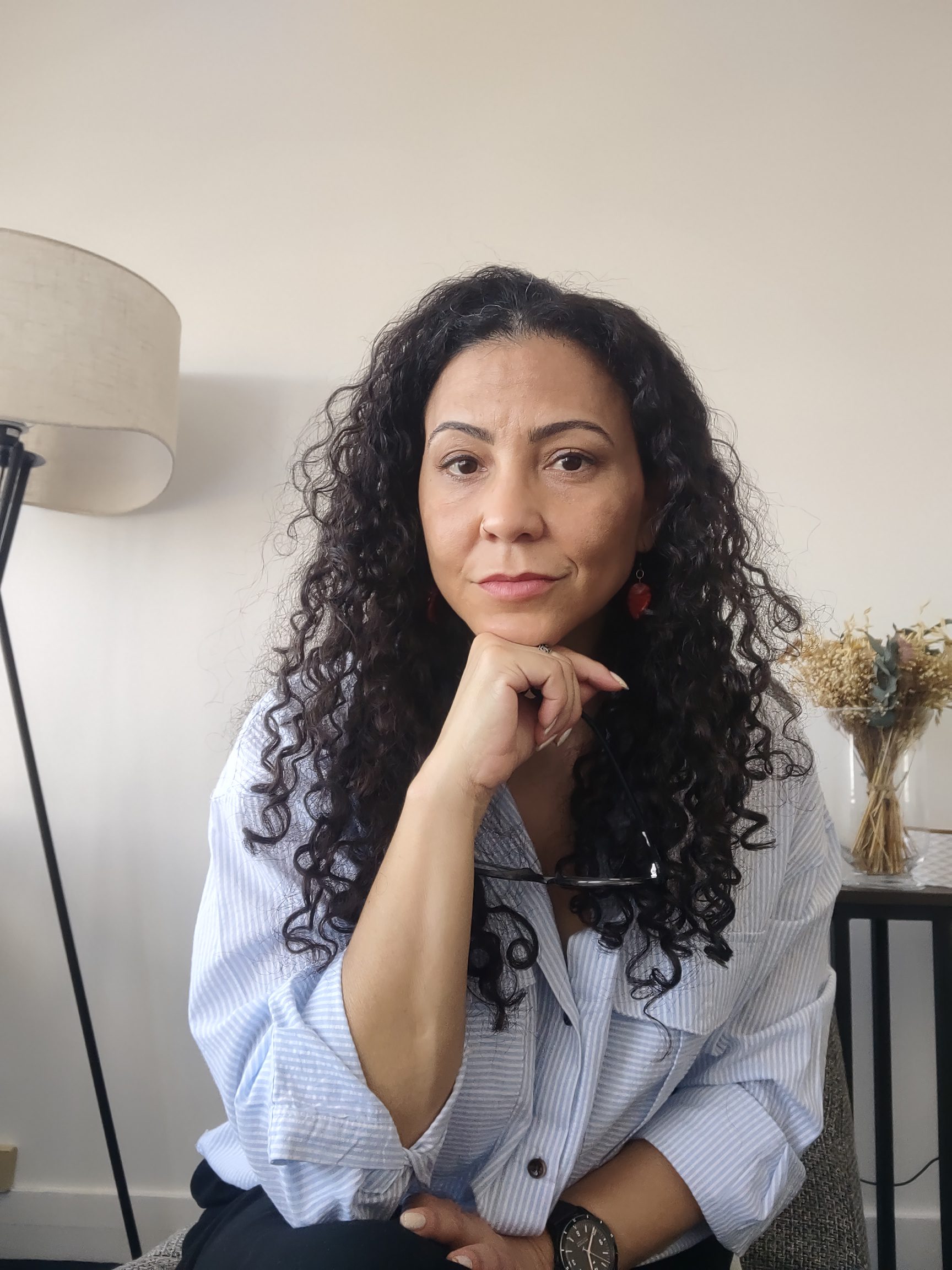Juliana Andrade, psicóloga clínica e do trabalho. Foto no consultório, sentada, vestindo camisa azul clara de manga.